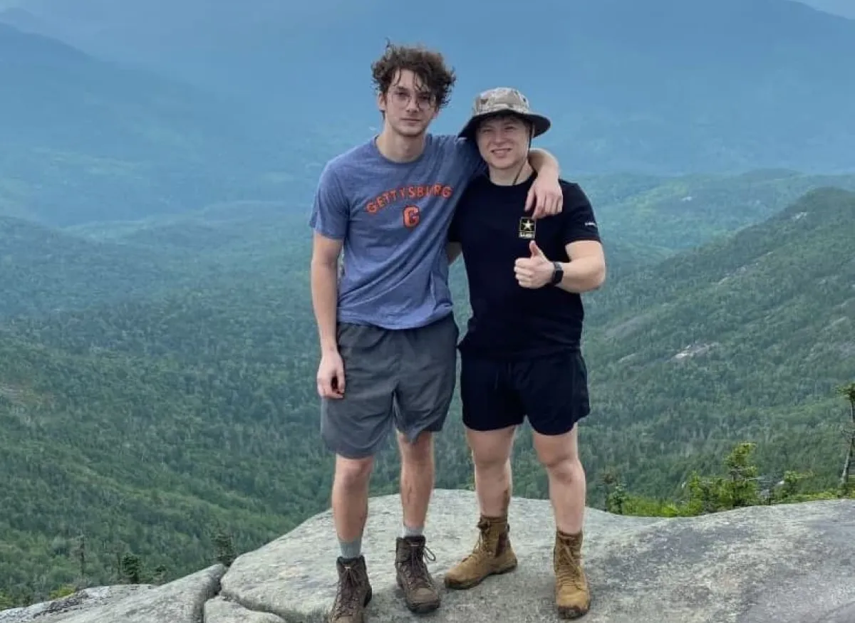 Two people standing together on a mountain overlook with forested hills and blue mountains in the background