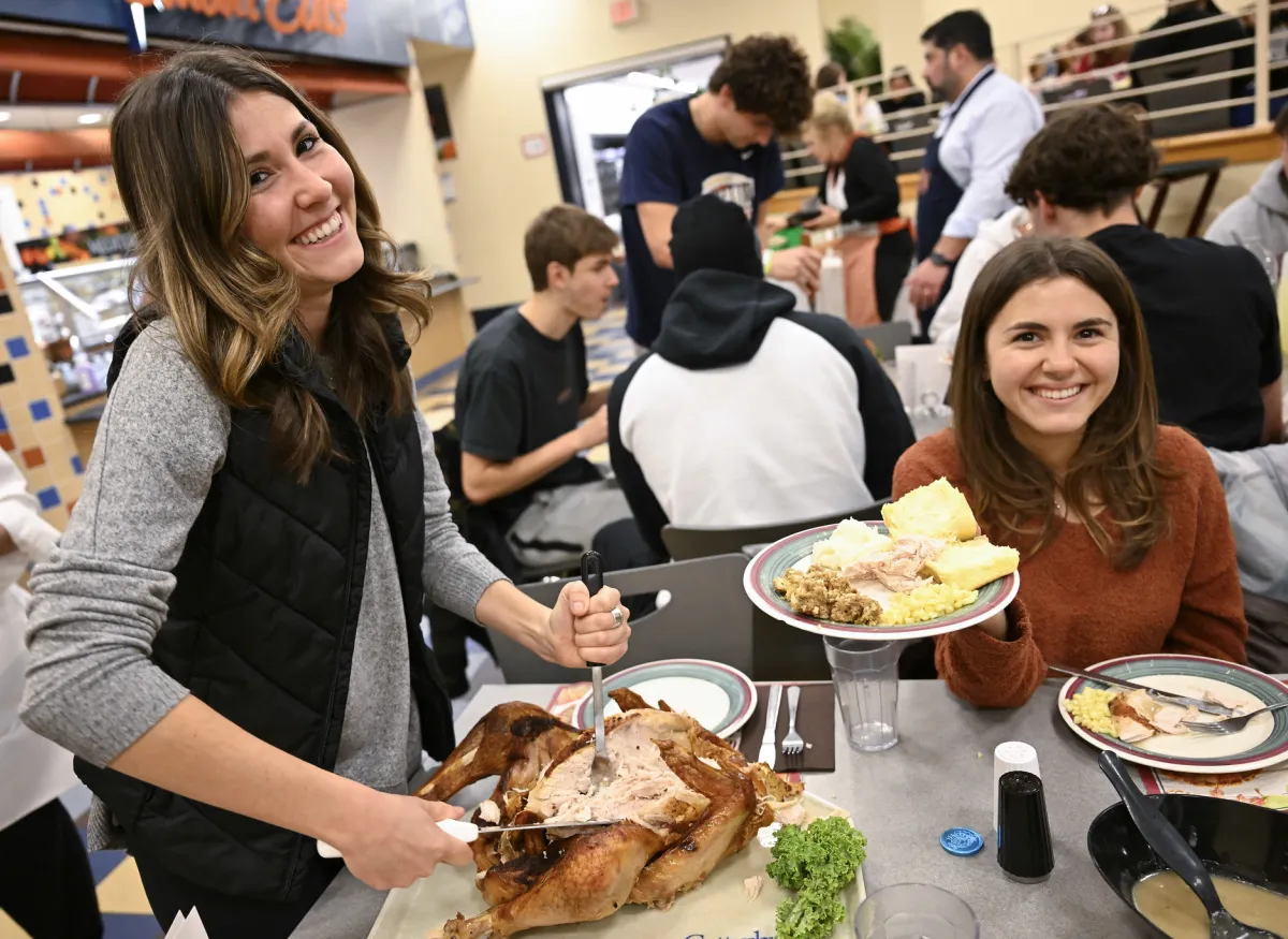 2 Students at Servo's Thankgivining dinner with a plate of food consisting of Turkey, corn, mashed potatoes and rolls