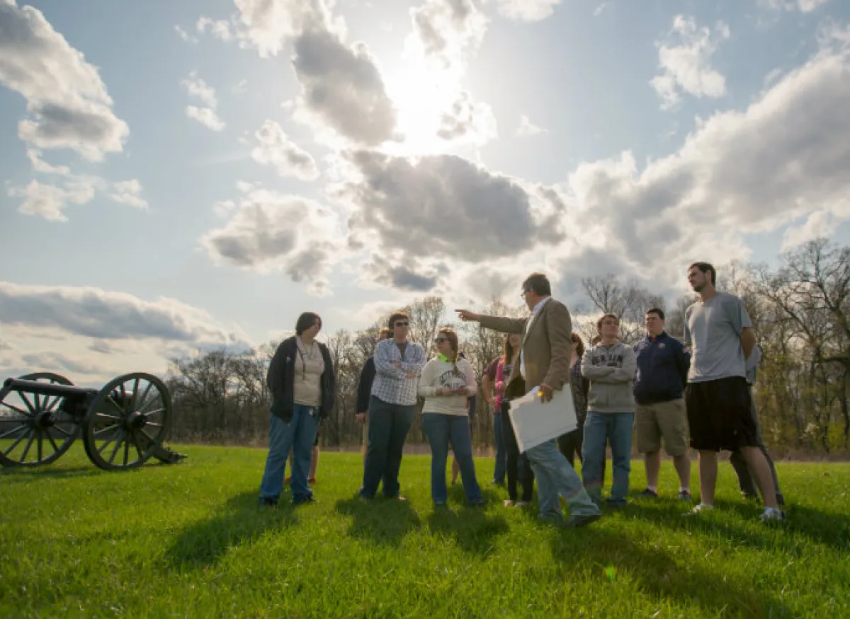 A professor and students stand on a grassy field beside a historic cannon
