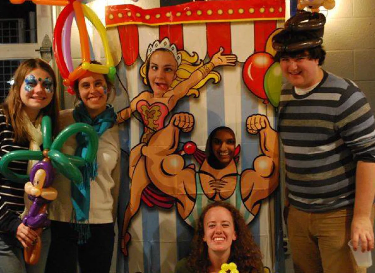 A group of people in colorful costumes poses indoors beside a circus-themed mural.
