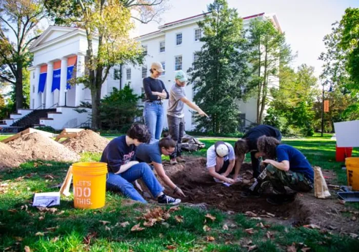 A group of people works together outdoors in a landscaped area, planting or tending to a garden bed near a large white building.
