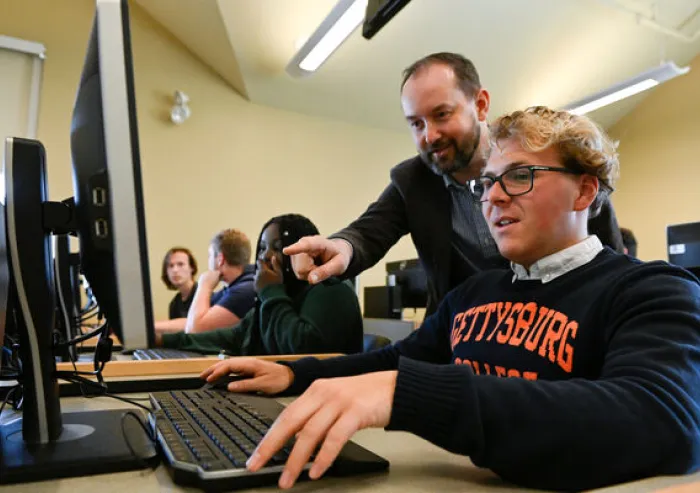 A student works at a computer while an instructor provides guidance in a classroom.
