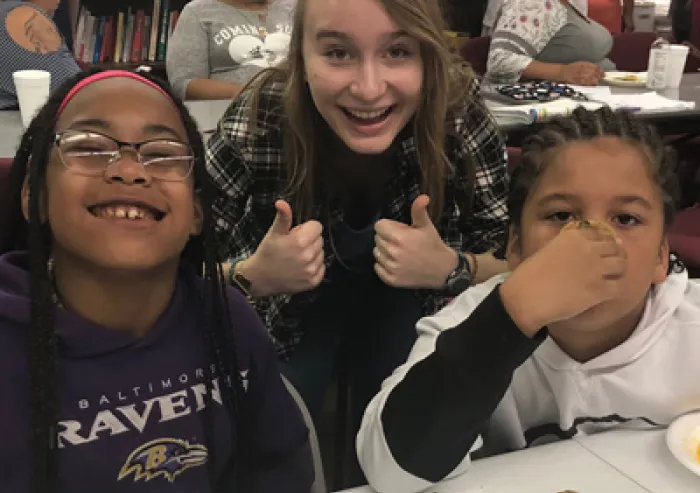 A student stands behind two small children while giving a double thumbs-up.