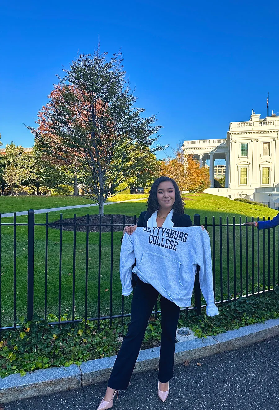 female student poses in front of White house holding up Gettysburg College sweatshirt