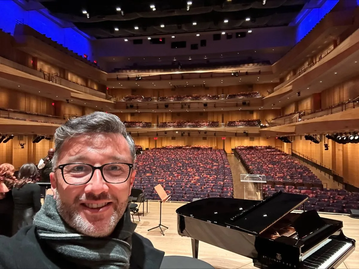 selfie of male prof on stage with students over one shoulder and piano and empty theater over other