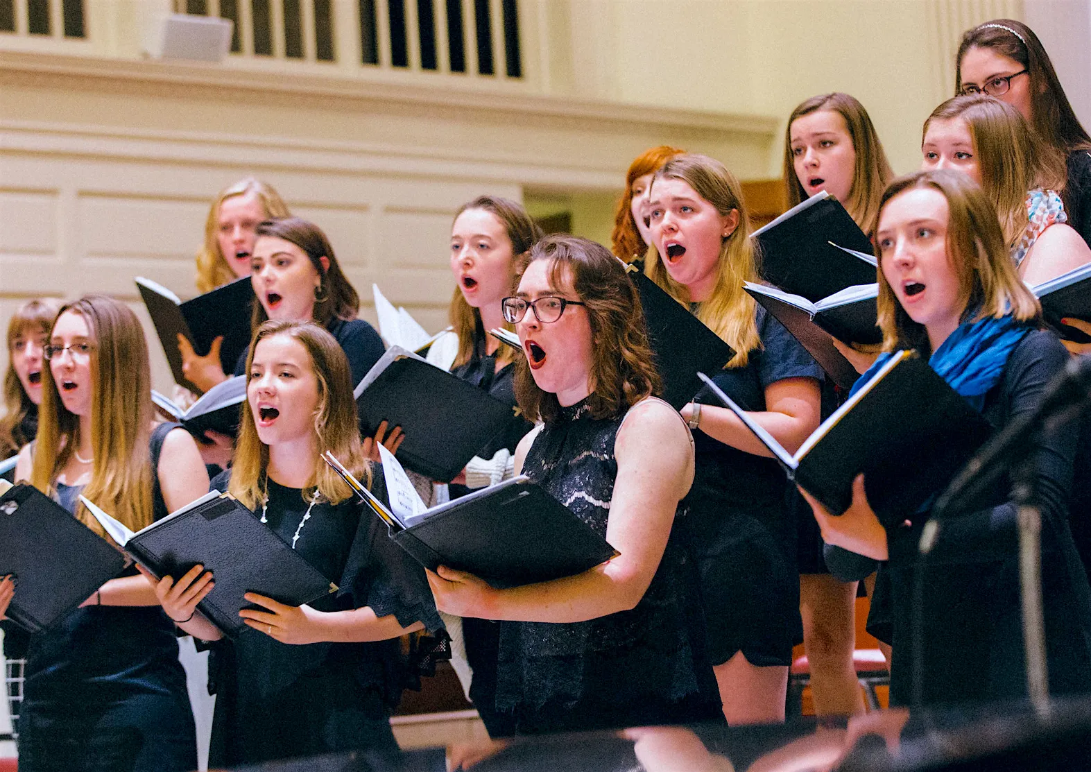 A choir performs while holding open sheet music, with singers dressed mostly in black in a formal interior.