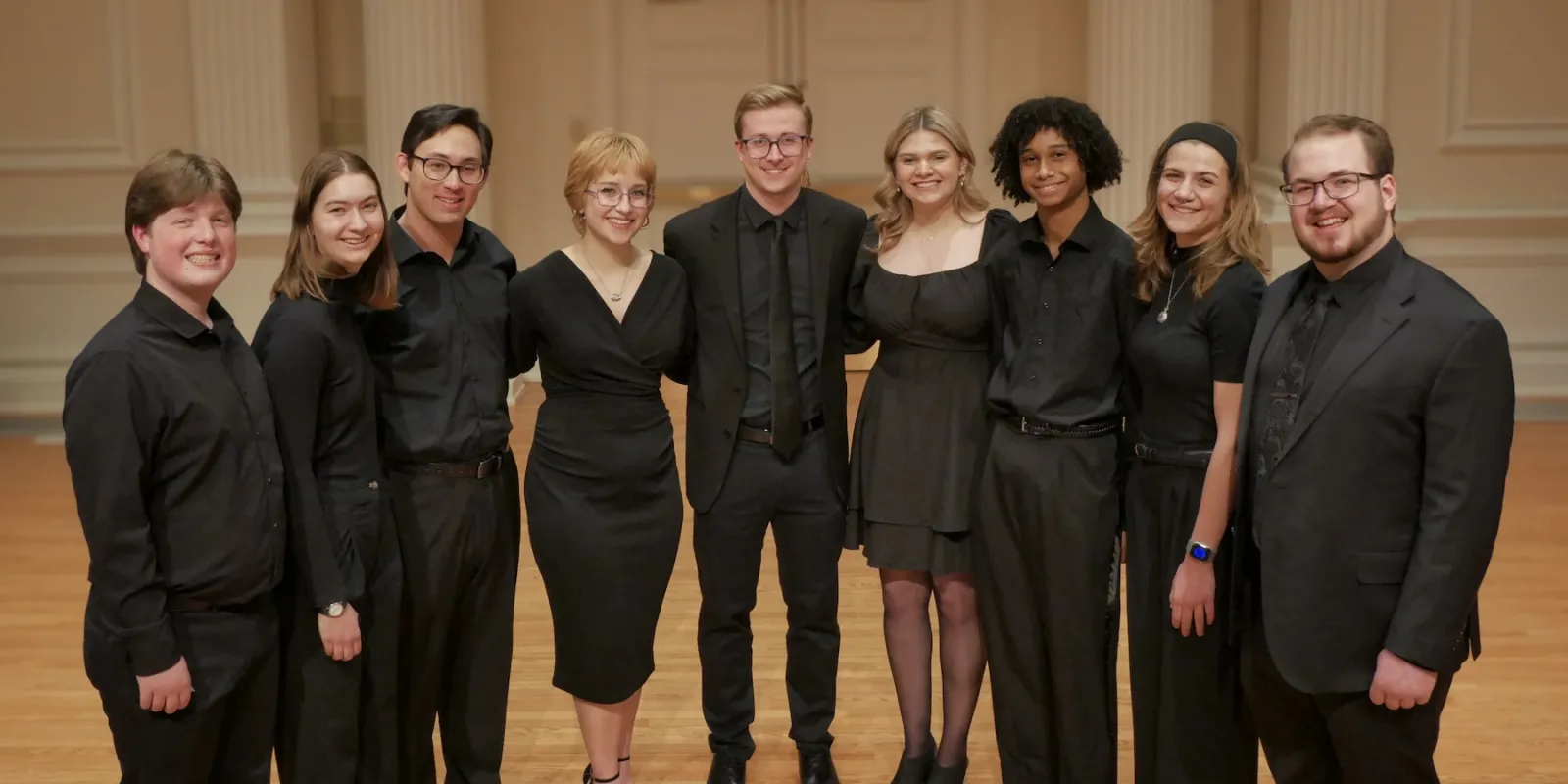 A group of nine performers poses together in a concert hall, all dressed in black.