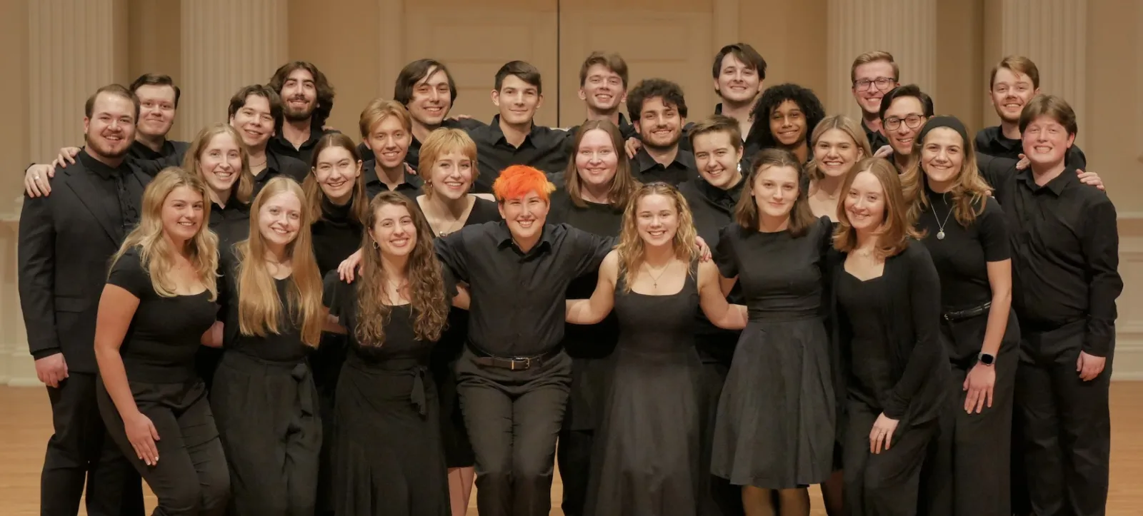 A large vocal ensemble poses together in a concert hall, with the members wearing coordinated black attire.