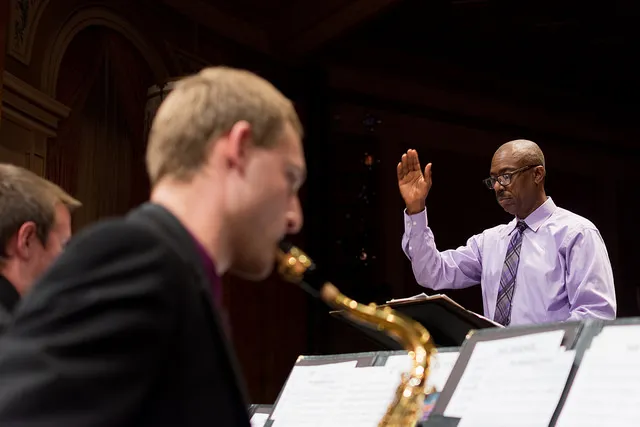 A jazz big band performs onstage with a conductor, featuring piano, guitar, saxophones, brass instruments, drums, and double bass.