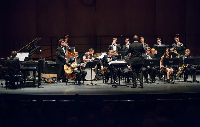 A jazz big band performs onstage with a conductor, featuring a pianist, guitarist, saxophones, trumpets, trombones, drums, and double bass.