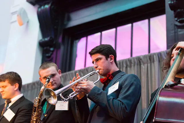 A small jazz ensemble performs onstage, featuring a trumpet player, saxophonist, and double bassist in formal attire.