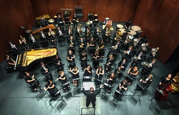 An overhead view shows a conductor leading a large concert band, including woodwinds, brass, percussion, piano, and double bass.