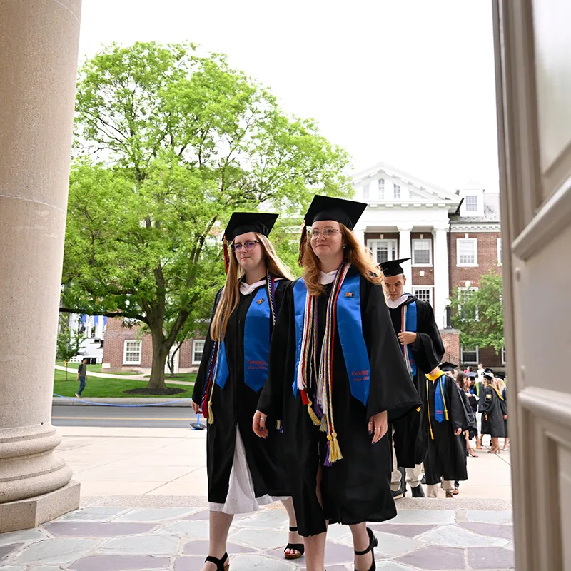 Graduates in black gowns and colorful stoles walk through a campus building.