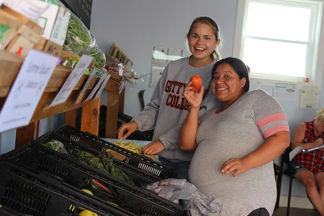 A smiling student stands next to a woman holding up a piece of fruit.