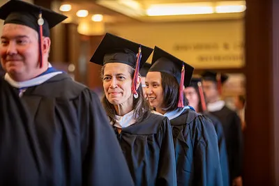 Graduates in black caps and gowns walk in a procession through an indoor venue. 