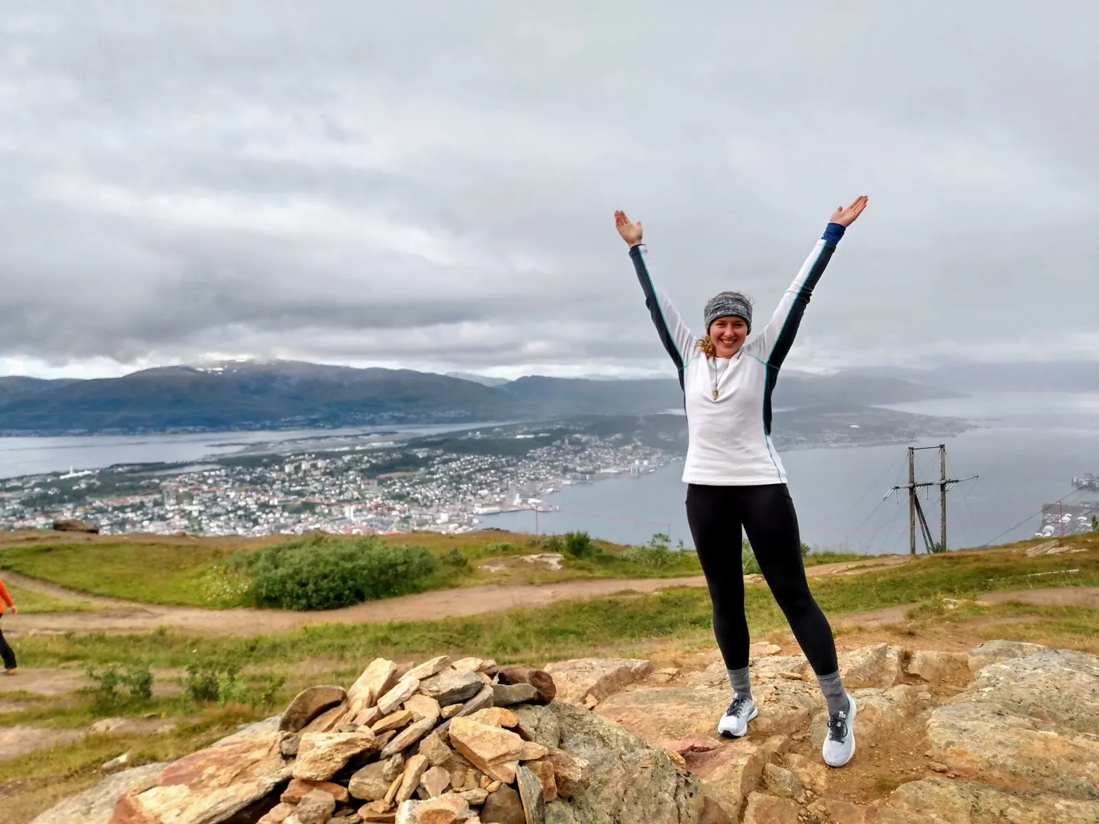 A person raises both arms while standing on a rocky overlook above a coastal city and mountain landscape.