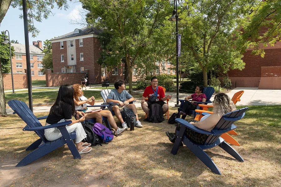 Students sit together in Adirondack chairs on a campus lawn, talking and spending time outdoors.