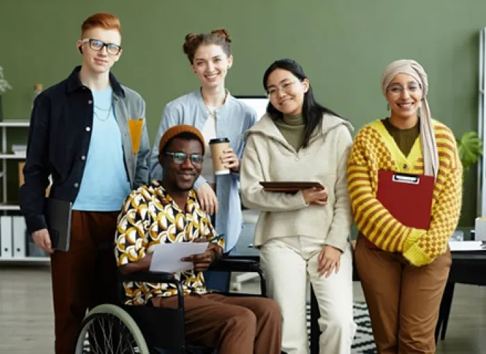Group of five students, including one student using a wheelchair, smiling together in a classroom.