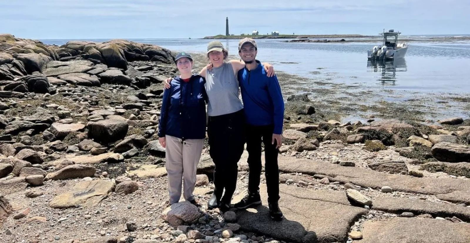 students pose on rocky beach with ocean, boat, and lighthouse in background