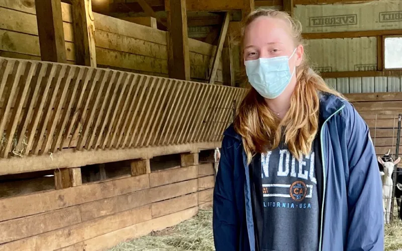masked female student stands in barn with goats in backround